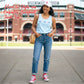 Woman wearing a light blue tank top with baseball and bow icons embroidered across the chest, jeans, and red sneakers in front of a stadium.