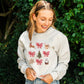 woman wearing a heather oatmeal pullover with a retro reindeer, bow, cocoa, tree, cookie, and snow globe printed design