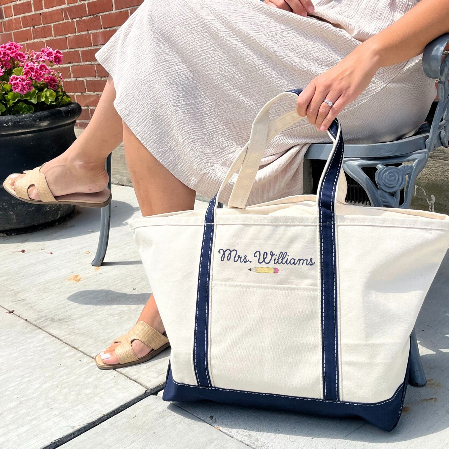 woman sitting on a bench outside with a large canvas tote bag with navy trim and handles, and 'Mrs. Williams' along with a mini pencil embroidered above the front outside pocket