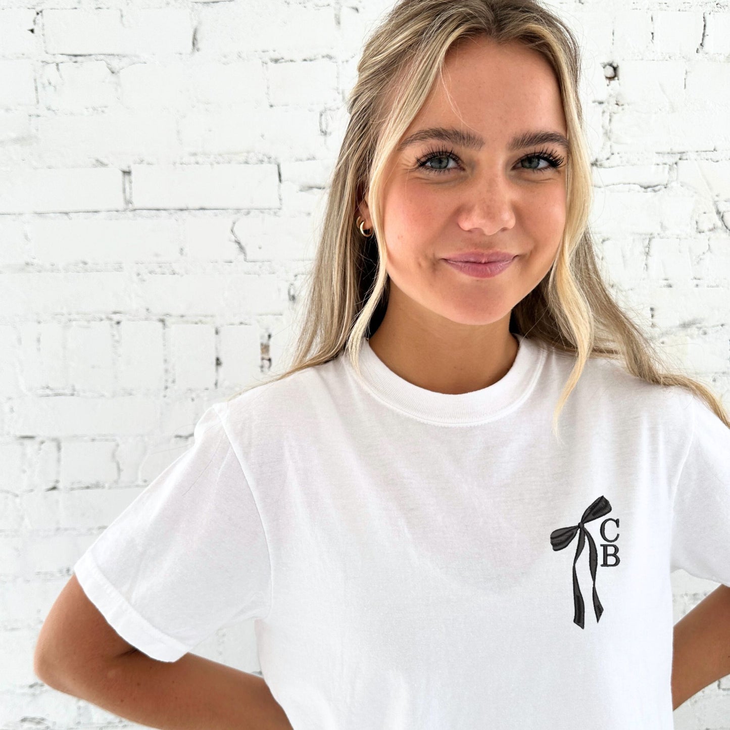 girl wearing a white comfort colors t-shirt with custom ribbon bow stacked initial embordered design on the left chest in black thread.