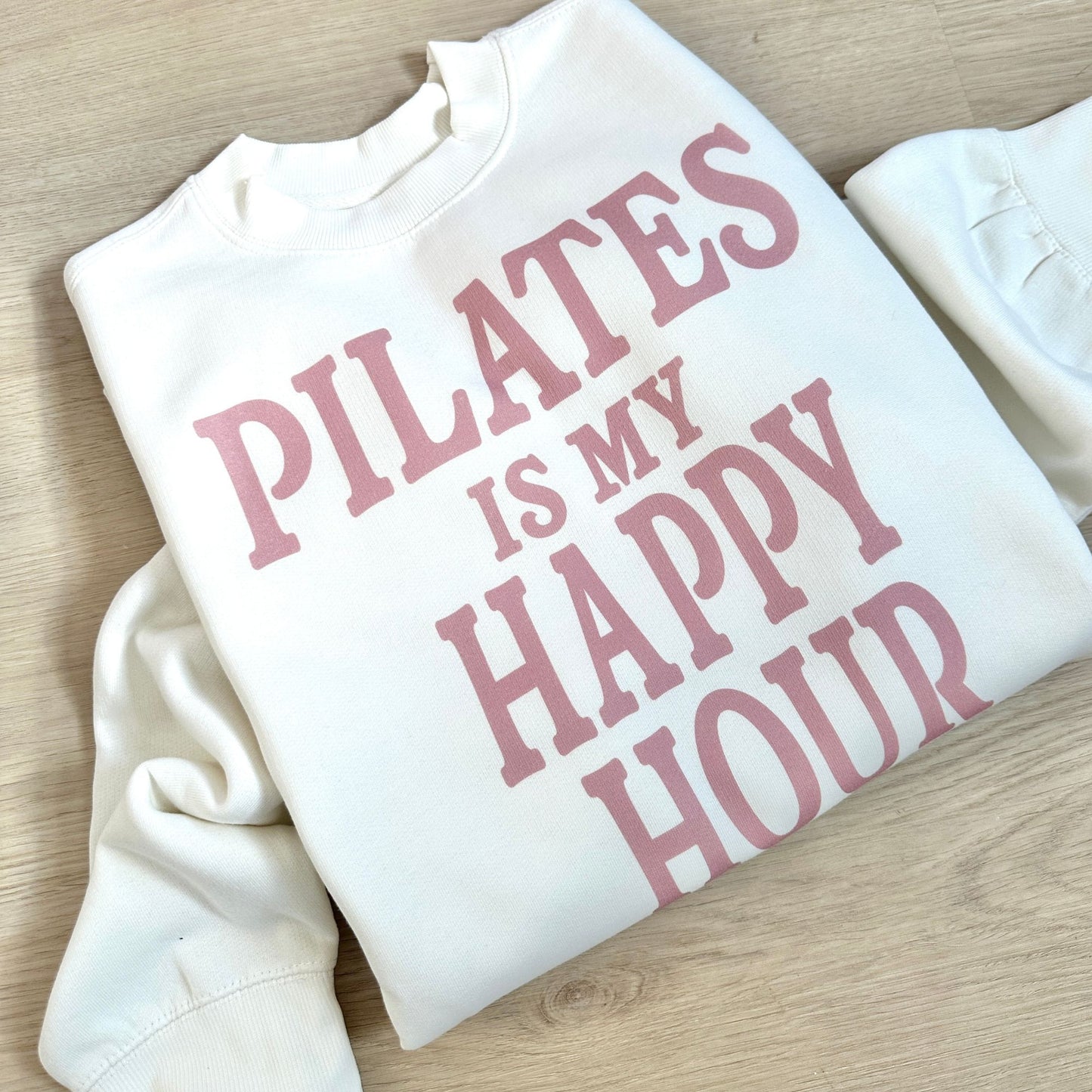 White sweatshirt with pink text 'PILATES IS MY HAPPY HOUR' on a wooden surface