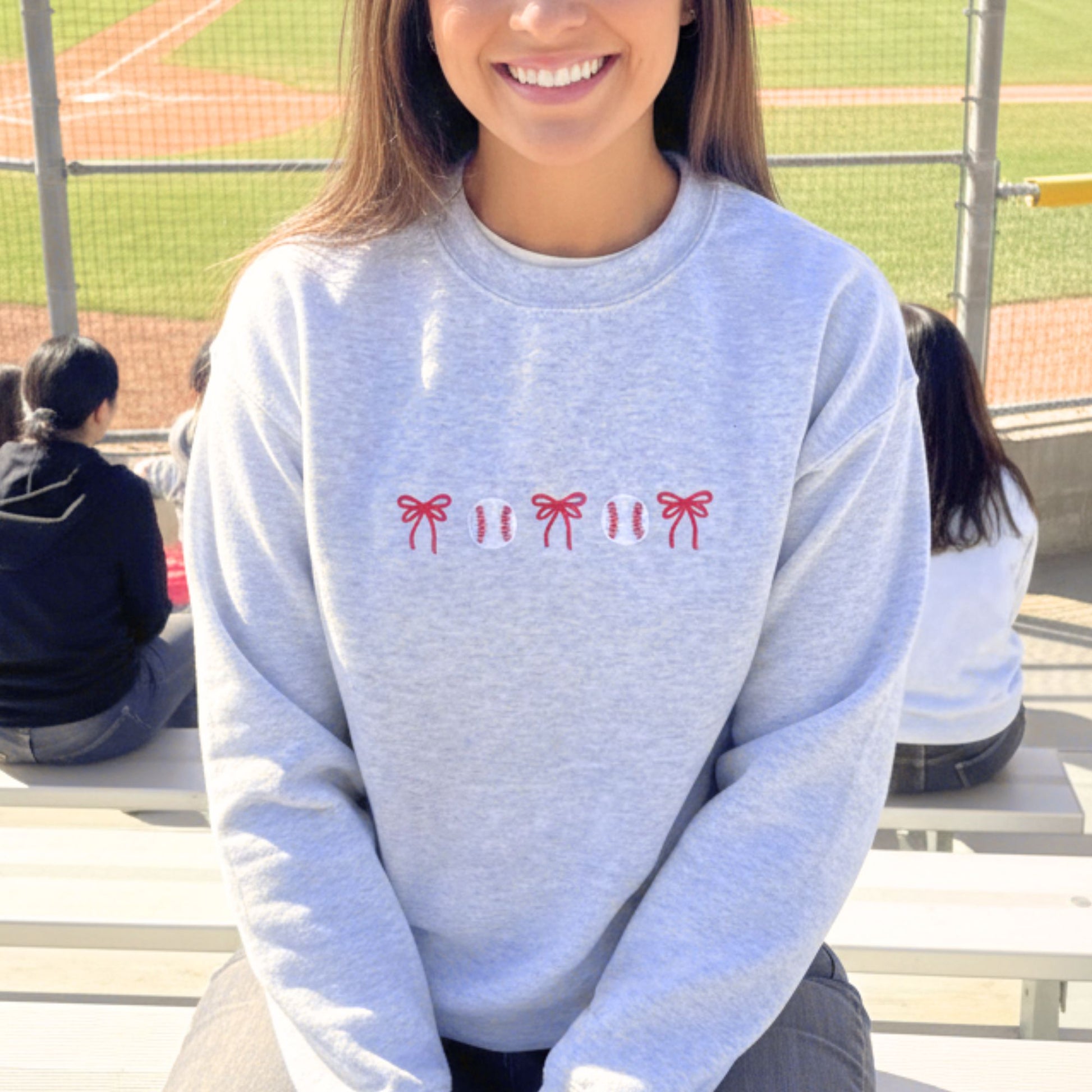 Person wearing an ash crewneck sweatshirt with red bow and baseball designs on a sports field background 