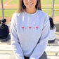 Person wearing an ash crewneck sweatshirt with red bow and baseball designs on a sports field background 