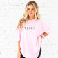 Woman wearing a pink t-shirt with text and graphics against a white brick wall.