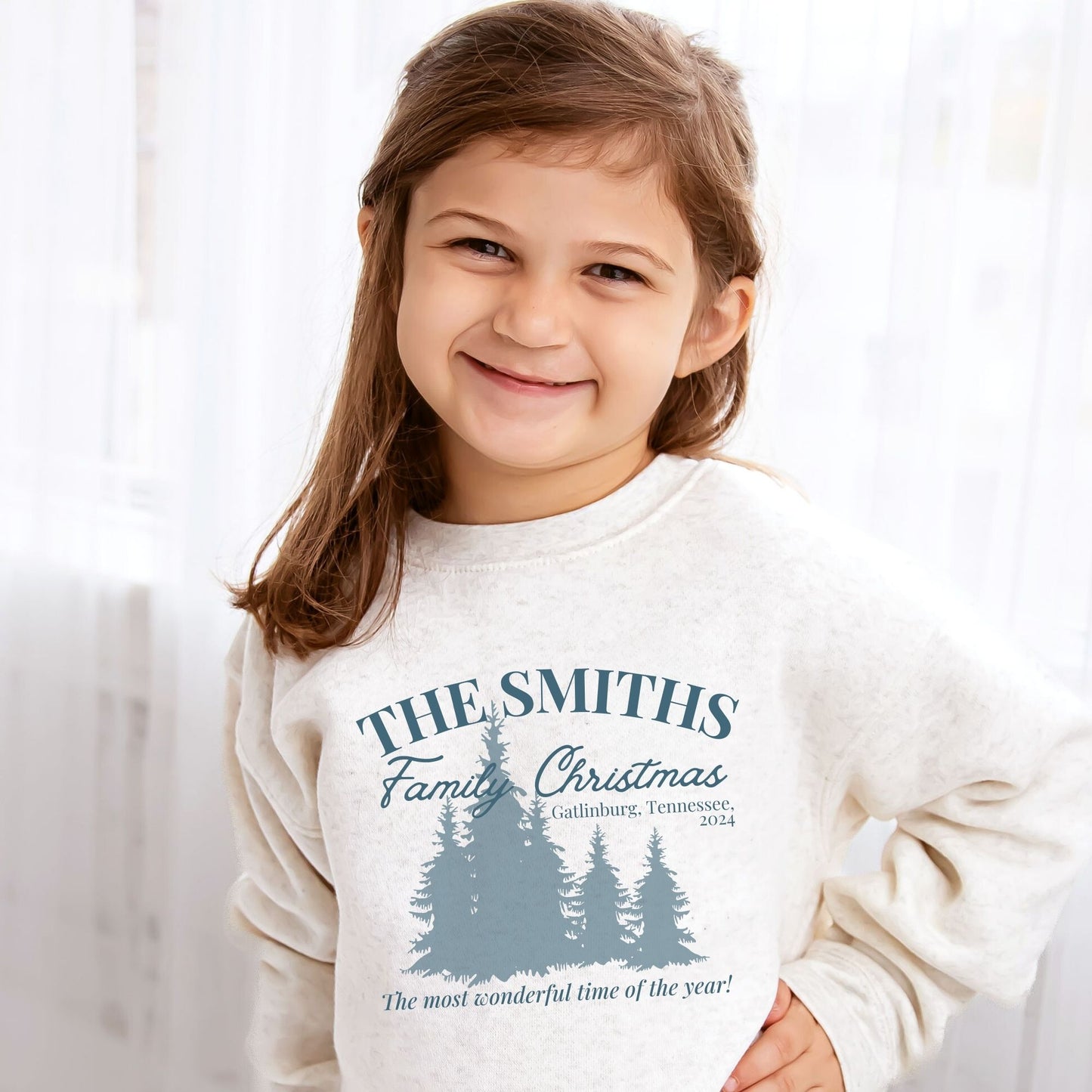 young girl wearing a natural heather youth sweatshirt with a custom christmas print featuring a family name, vacation location, year, trees, and a "The most wonderful time of the year!" quote in blue ink