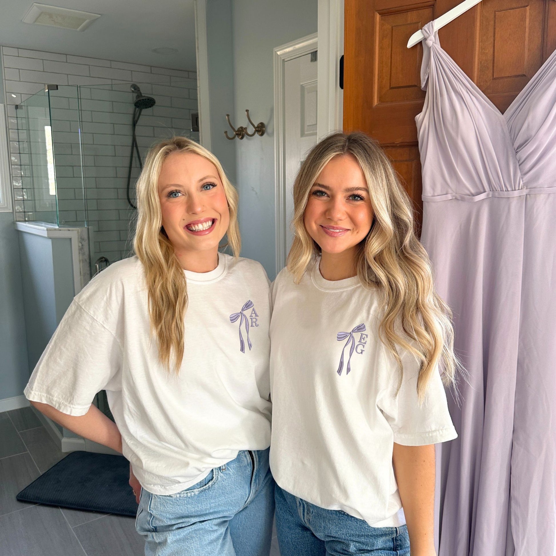 Two girls wearing white comfort colors t-shirts with custom ribbon bow and stacked initial embroidery on the left chest in smoky orchid thread.