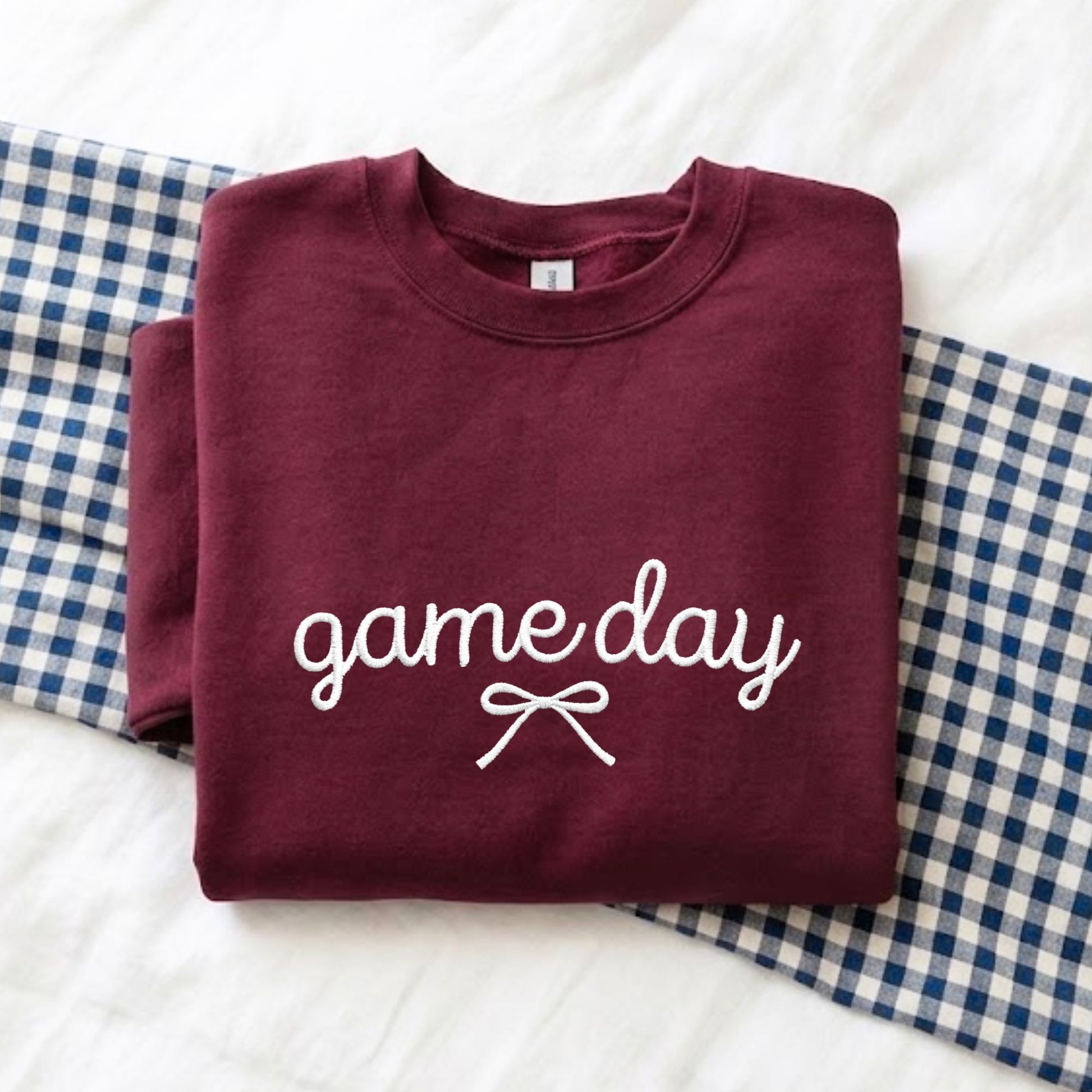 A folded maroon crewneck sweatshirt with 'game day' and a bow embroidered design in white on the front, displayed on a white textured blanket with blue-and-white gingham fabric underneath.