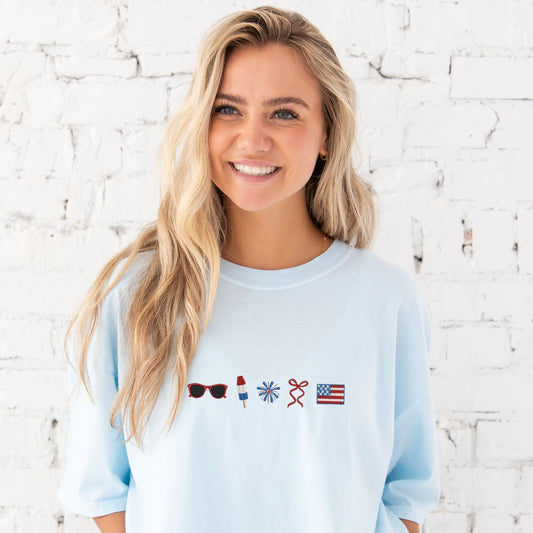 girl standing in front of a white brick wall wearing an oversized comfort colors chambray blue t-shirt with 4th of July inspired embroidered icons: red sunglasses, a red white and blue popsicle, firework, red bow, and an American flag.