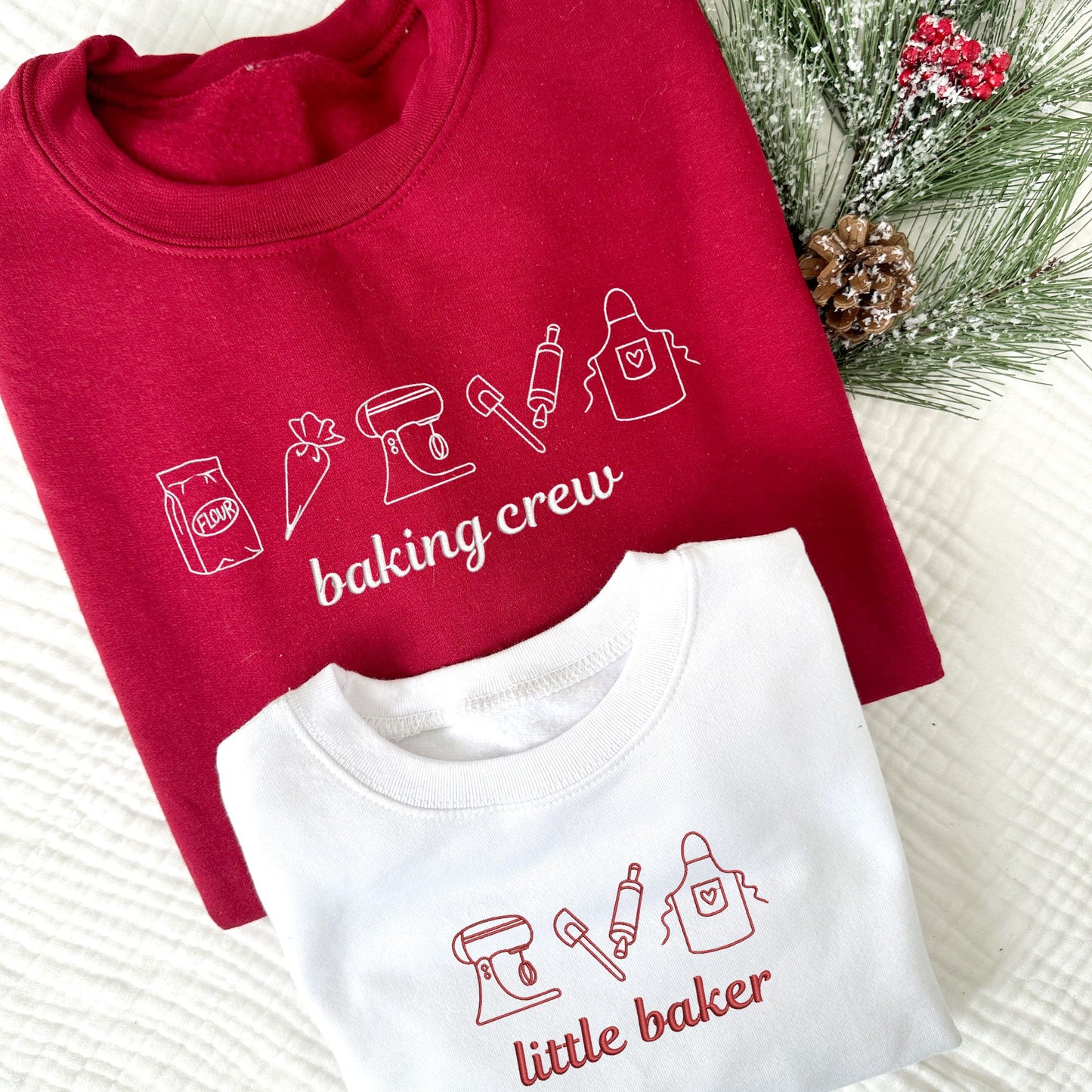 Adult cardinal red crewneck with embroidered baking icons and baking crew in white thread. Youth white crewneck with embroidered baking icons and little baker in red thread.