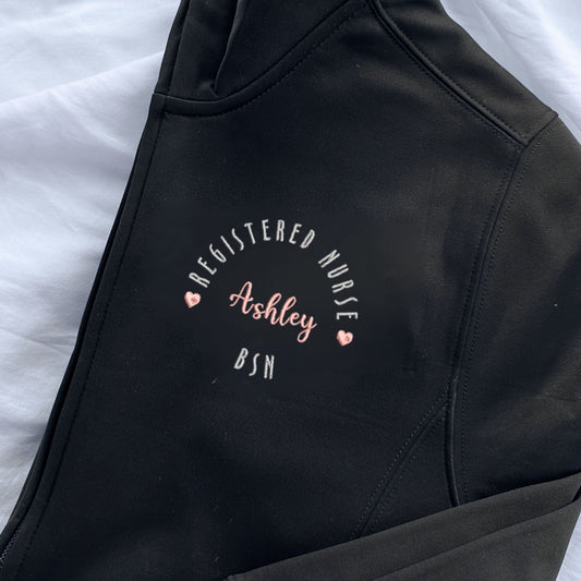 a black polyester full zip jacket with custom embroidered registered nurse design on the left chest featuring hearts, name, and credentials.