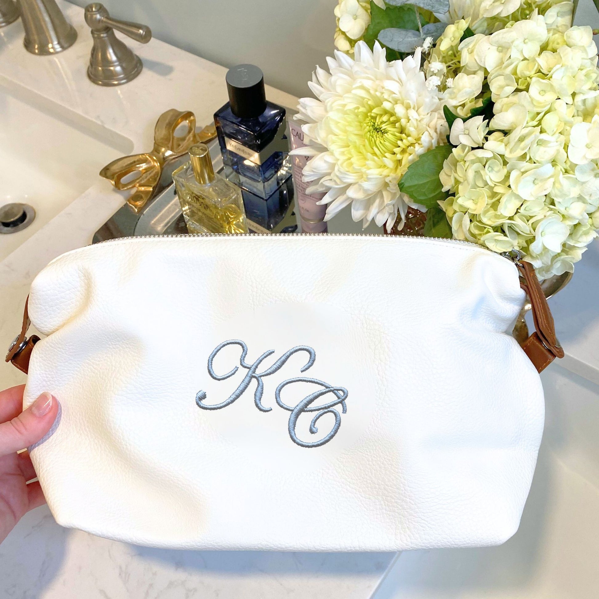 White pouch with 'KC' monogram, flowers, and perfume bottles on a bathroom counter.