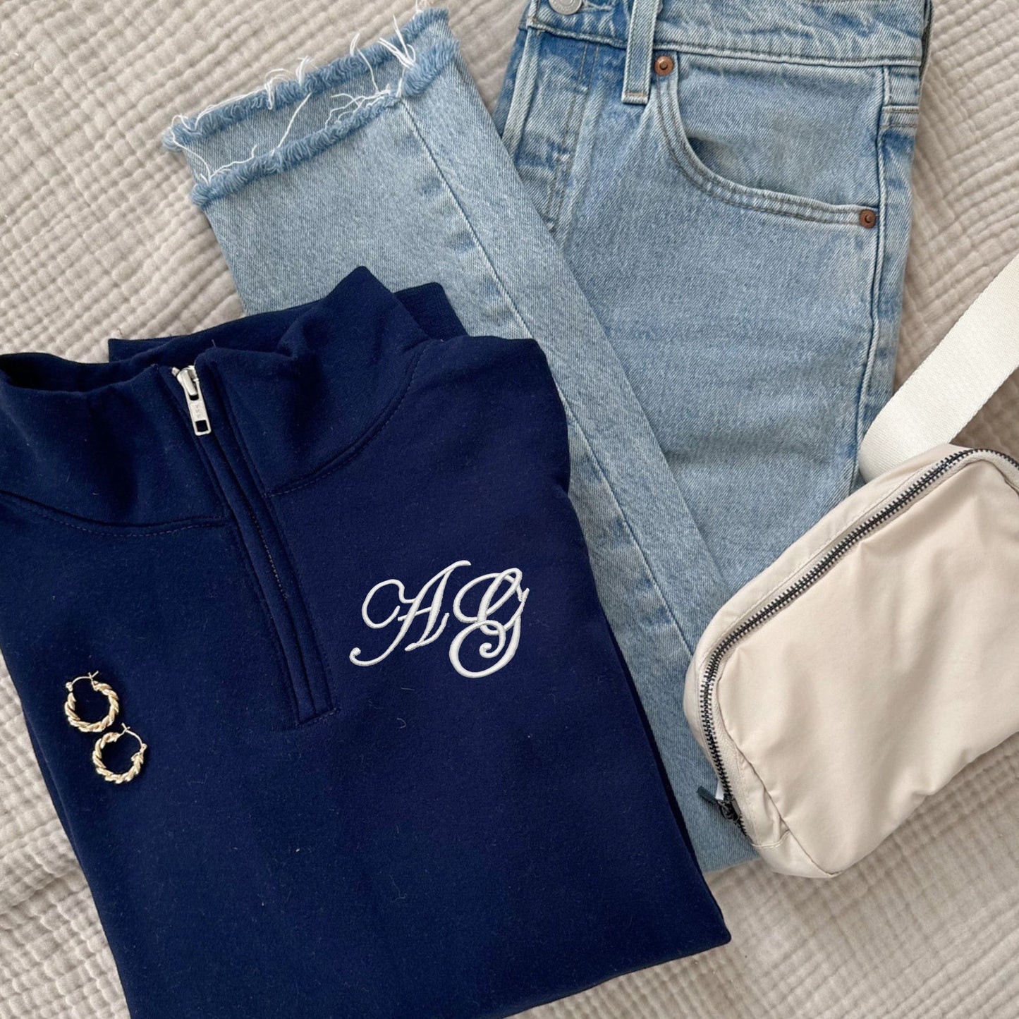 Navy blue quarter zip sweatshirt with edwardian script 2 letter monogram in white thread, light blue jeans, gold hoop earrings, and beige pouch on a textured surface.