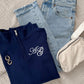Navy blue quarter zip sweatshirt with edwardian script 2 letter monogram in white thread, light blue jeans, gold hoop earrings, and beige pouch on a textured surface.