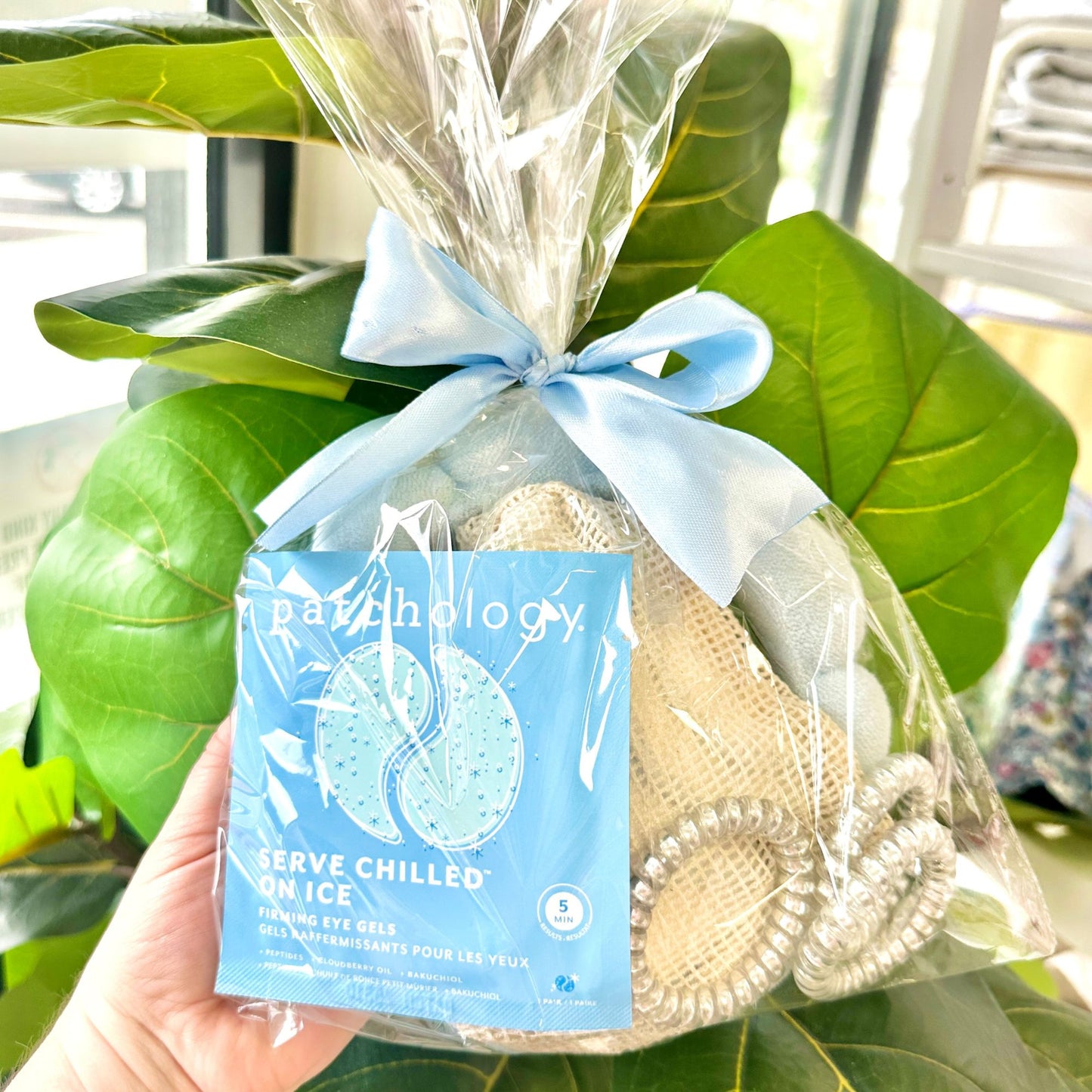 Packaged eye gels, reusable cotton rounds, spa head band, and coiled hair ties in shades of blue in a clear bag and tied with a decorative bow held in front of green leaves