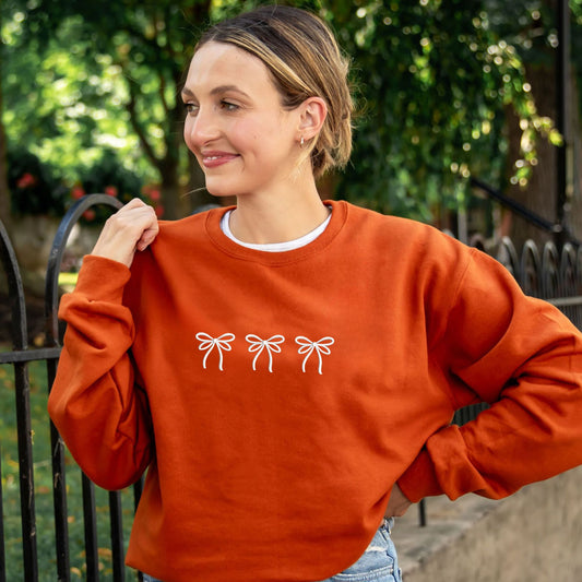 Woman wearing a Texas Orange crewneck sweatshirt with 3 white double bow ribbons embroidered across the chest in white thread.