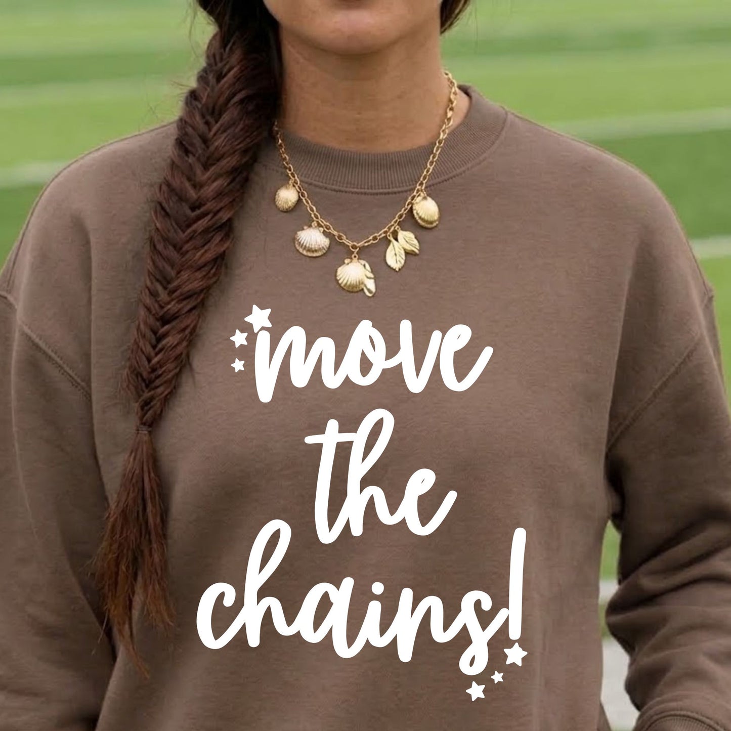 A person wearing a brown crewneck sweatshirt with the white text 'move the chains!' printed on the front, paired with a gold shell and leaf charm necklace, set against a blurred green athletic field background.
