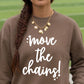 A person wearing a brown crewneck sweatshirt with the white text 'move the chains!' printed on the front, paired with a gold shell and leaf charm necklace, set against a blurred green athletic field background.