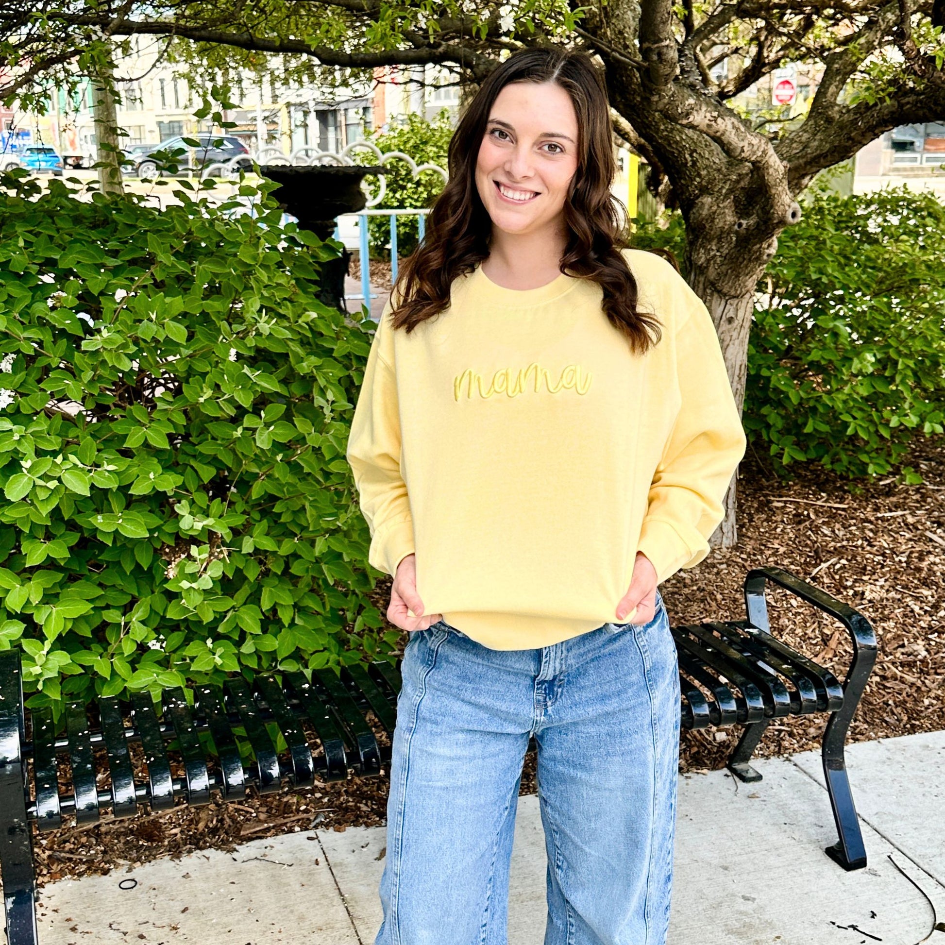 Woman wearing a yellow sweatshirt with text, standing outdoors near greenery and a bench.