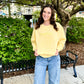 Woman wearing a yellow sweatshirt with text, standing outdoors near greenery and a bench.