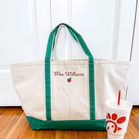 Canvas tote bag with green trim and handles, featuring 'Mrs. Williams' and an apple logo embroidered above the front outside pocket, sitting next to a white cup 
