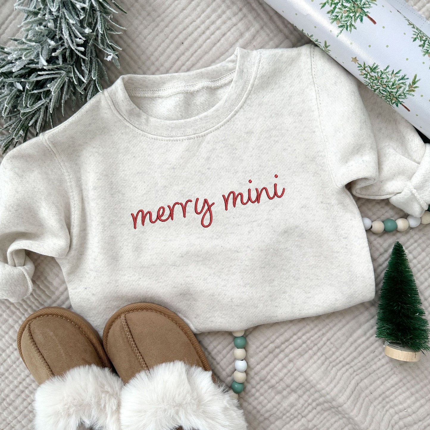 toddler heather oatmeal crewneck sweatshirt with embroidered merry mini design n red thread across the chest styled with Christmas décor