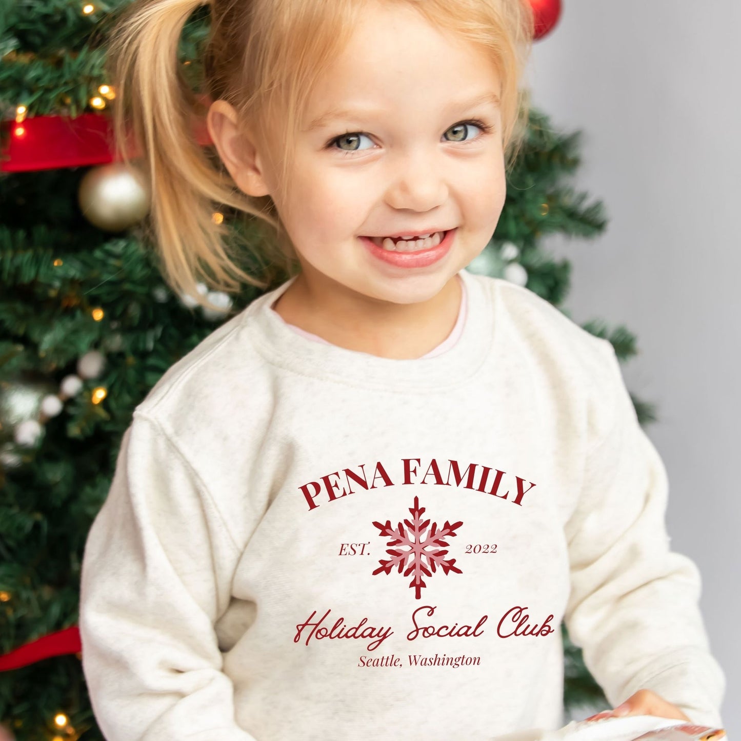 Little girl wearing a Natural Heather toddler crew with a custom print featuring a family name, Est date, snowflake, holiday social club, and location in red