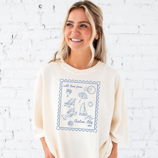 Woman standing in front of a white brick wall wearing an oversized ivory comfort colors t-shirt with a printed design of a large stamp with coastal inspired doodles inside of it and custom script text reading with love from... "custom city" xo" in blue ink