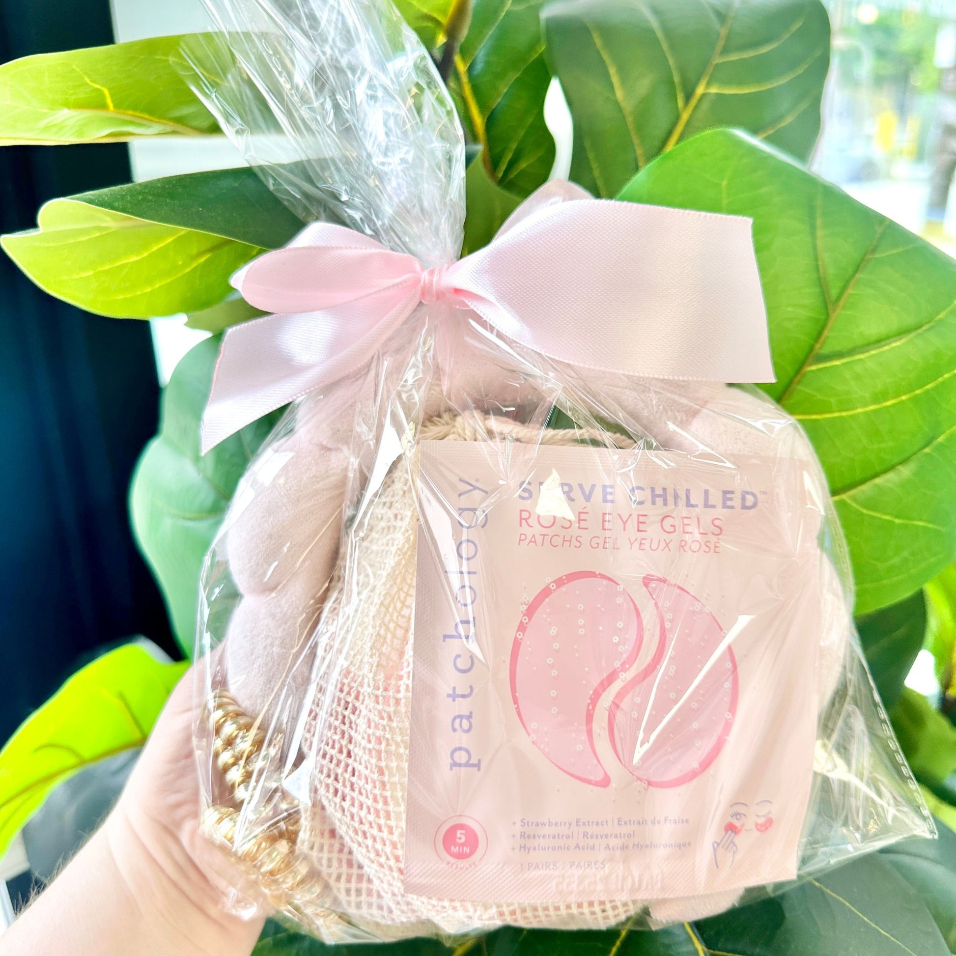 Packaged eye gels, reusable cotton rounds, spa head band, and coiled hair ties in shades of pink in a clear bag and tied with a decorative bow held in front of green leaves