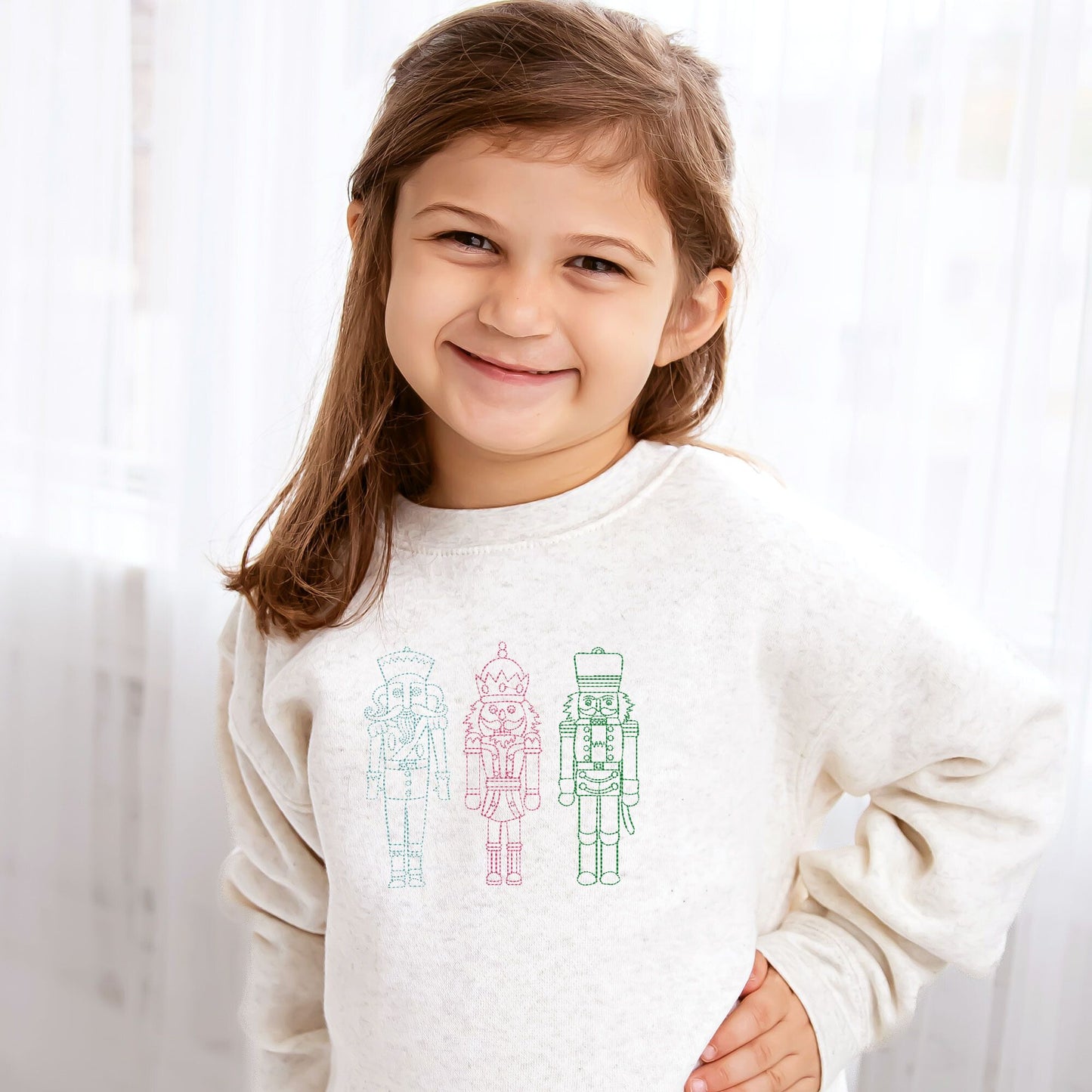 toddler girl wearing a heather oat crewneck sweatshirt with embroidered stitched nutcrackers across the chest in sky blue, pink, and kelly green