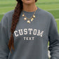 A close-up of a person wearing a slate gray crewneck sweatshirt with 'CUSTOM TEXT' embroidered in white arched letters on the chest. The person has a brown braid and is wearing a gold necklace, set against a blurred green athletic field background.