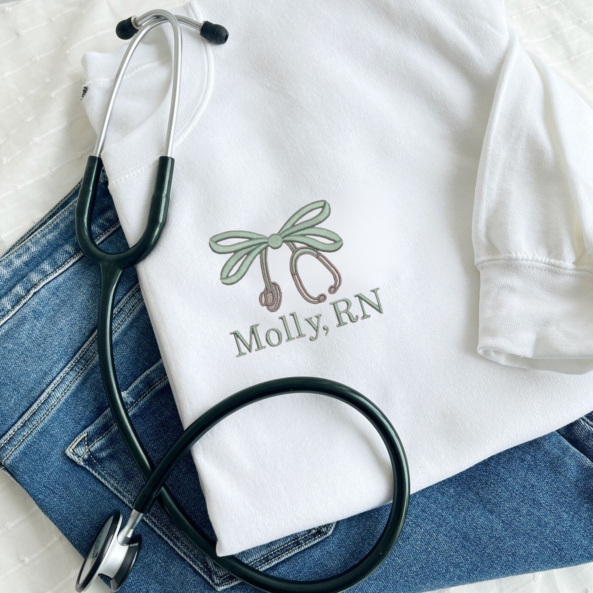 white crewneck sweatshirt with custom embroidered double loop bow stethoscope design and name on the left chest in eucalyptus and grey thread colors