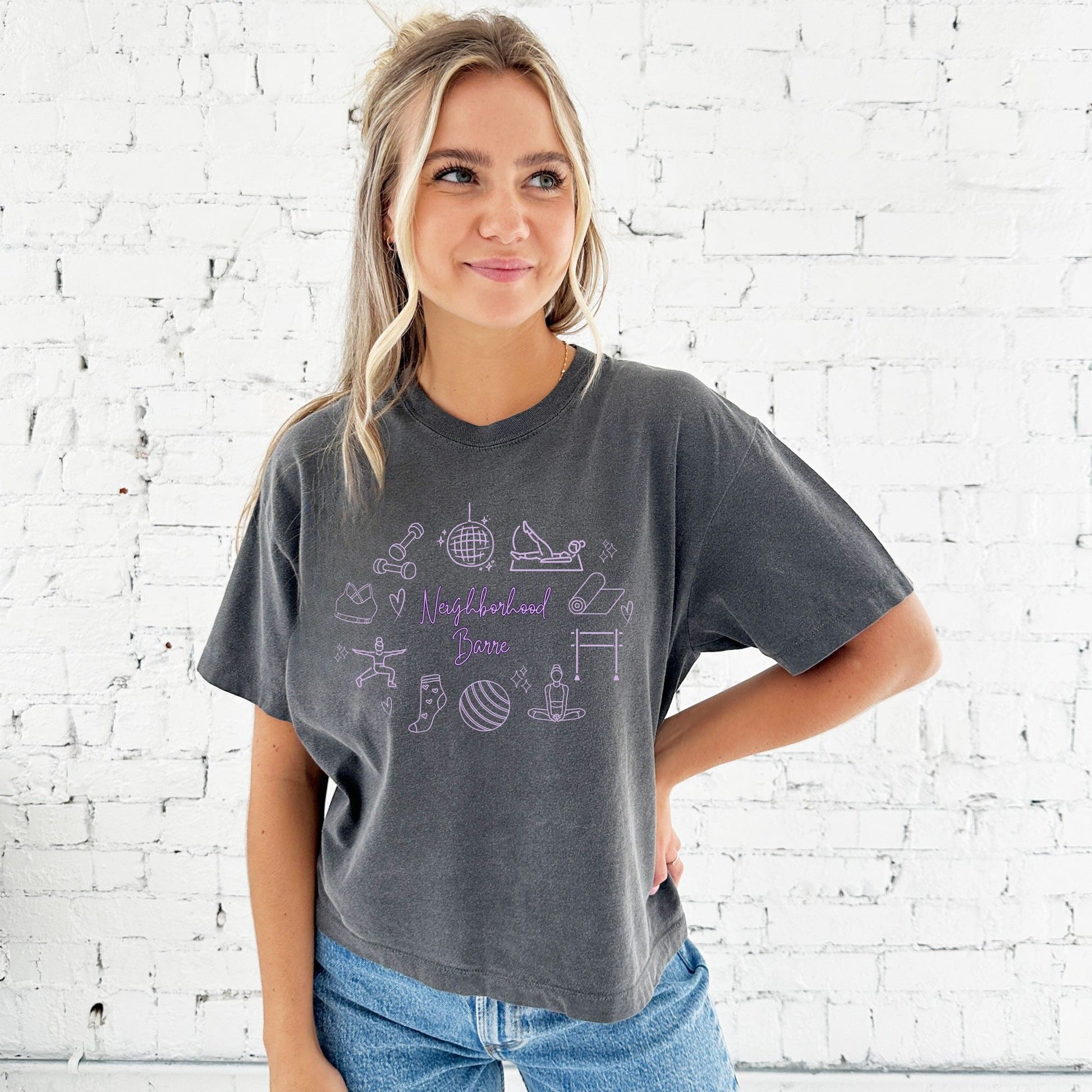 Woman wearing a gray t-shirt with purple graphics and text against a white brick wall.