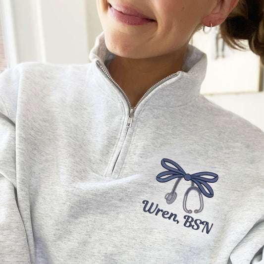 woman wearing an ash quarter zip sweatshirt with custom double loop bow stethoscope design with name embroidered on the left chest in navy thread