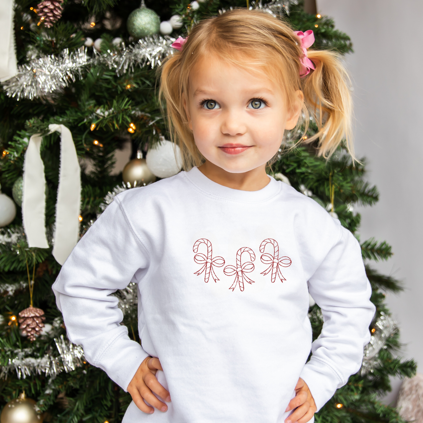 toddler girl wearing a white crewneck sweatshirt with 3 outlined candy canes wrapped in bows embroidered across the chest in red thread.