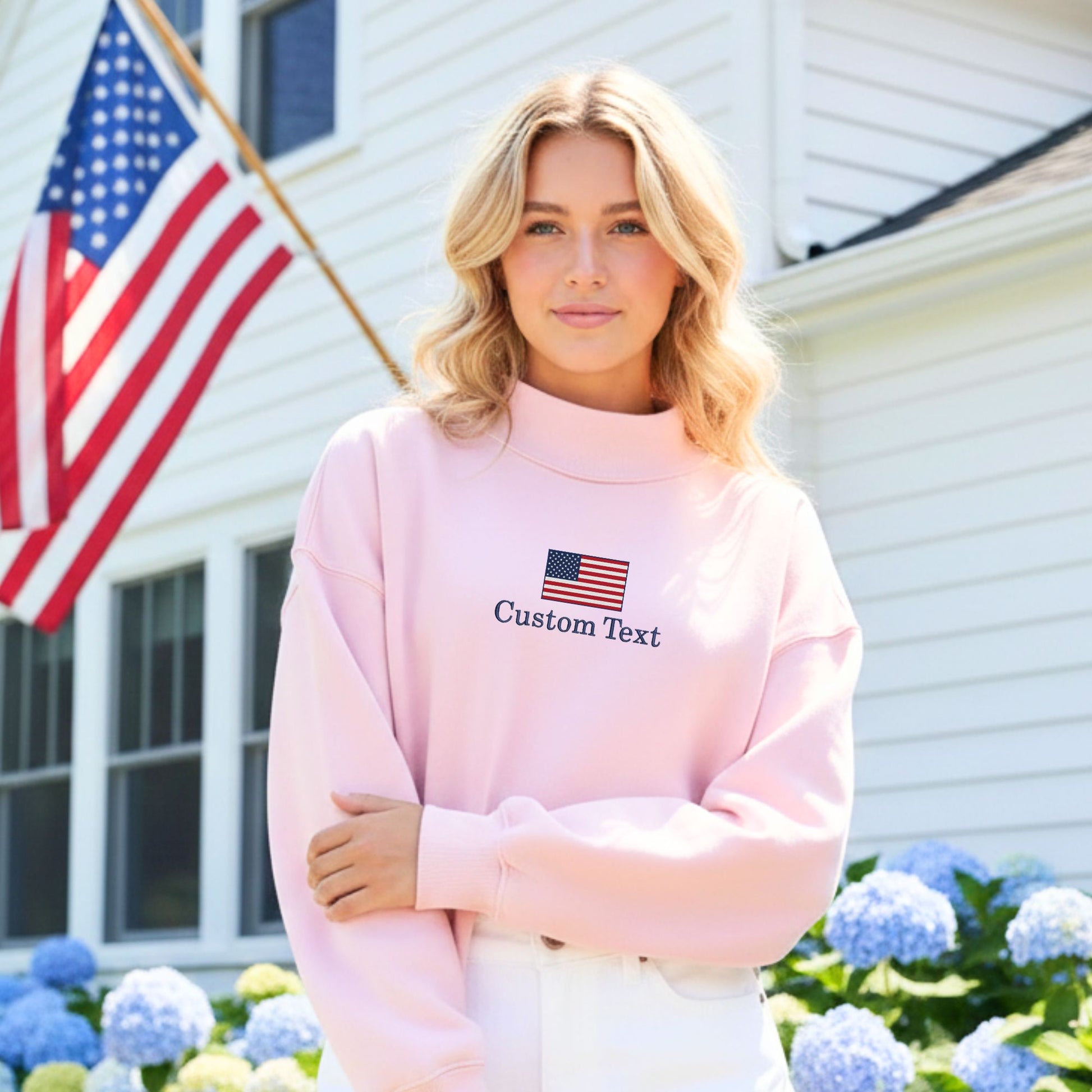 Woman wearing a pink sweatshirt with an American flag and text in front of a house with an American flag.