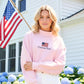 Woman wearing a pink sweatshirt with an American flag and text in front of a house with an American flag.