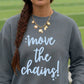 A person wearing a storm gray sweatshirt with the phrase 'move the chains!' printed in light blue script and small star accents. The person has a long brown braid and is wearing a gold necklace, photographed outdoors with a blurred green field in the background.