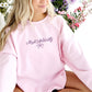 young woman wearing a light pink crewneck with embroidered Most Ardently and bow design in violet thread