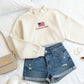Beige sweatshirt with American flag and text, paired with denim shorts on a white surface.