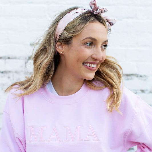 woman wearing a light pink crewneck sweatshirt with embroidered mama in baby pink thread an in all caps across the chest