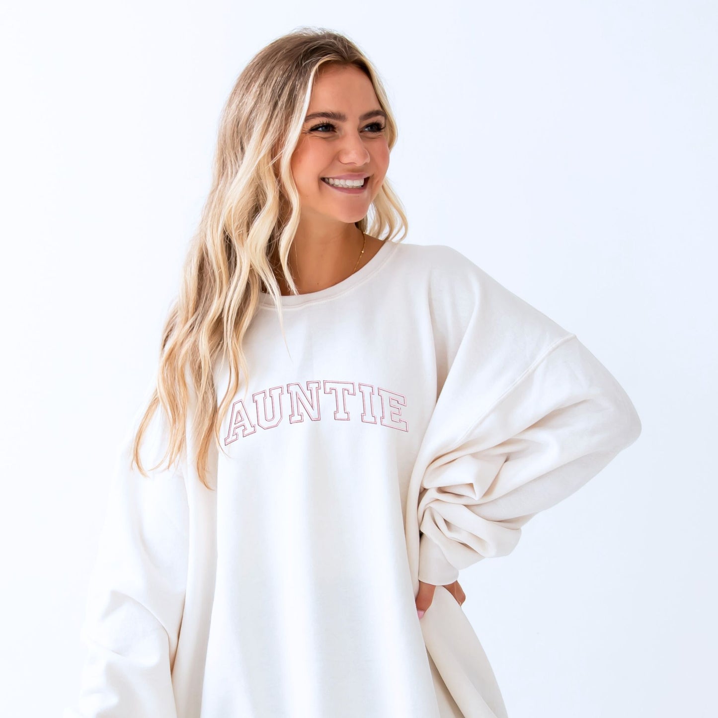 young woman wearing an oversized white crewneck sweatshirt with embroidered AUNTIE in all caps athletic block font across the chest in baby pink thread