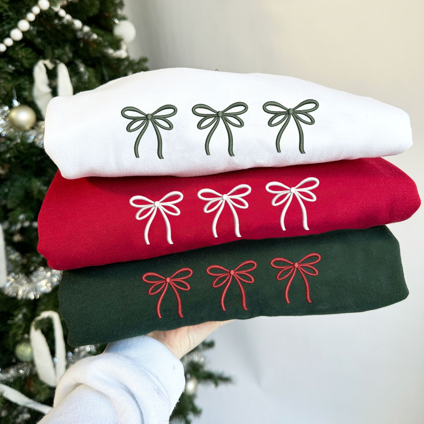 Christmas styled photo on someone holding up three crewneck sweatshirts in white, cardinal red and forest green. embroidered on the sweatshirt are three dainty ribbon bows in ivy, white, and red threads. 