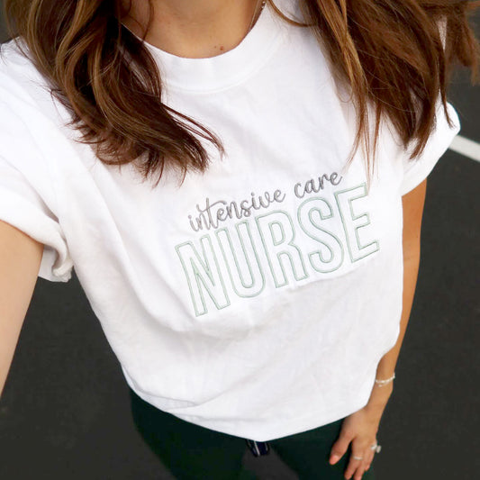 nurse wearing green figs scrub pants and a white comfort colors t-shirt with a custom intensive care nurse embroidered design in gray and eucalyptus