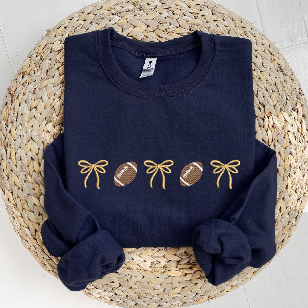 flat lay of a navy crewneck sweatshirt with embroidered alternating bows and footballs design across the chest. The bow are embroidered in gold thread.