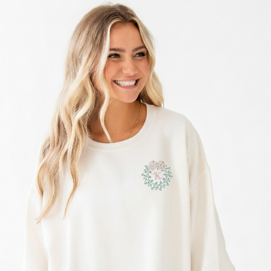 Woman wearing a white sweatshirt with a small design wreath bow initial design embroidered on the left chest 