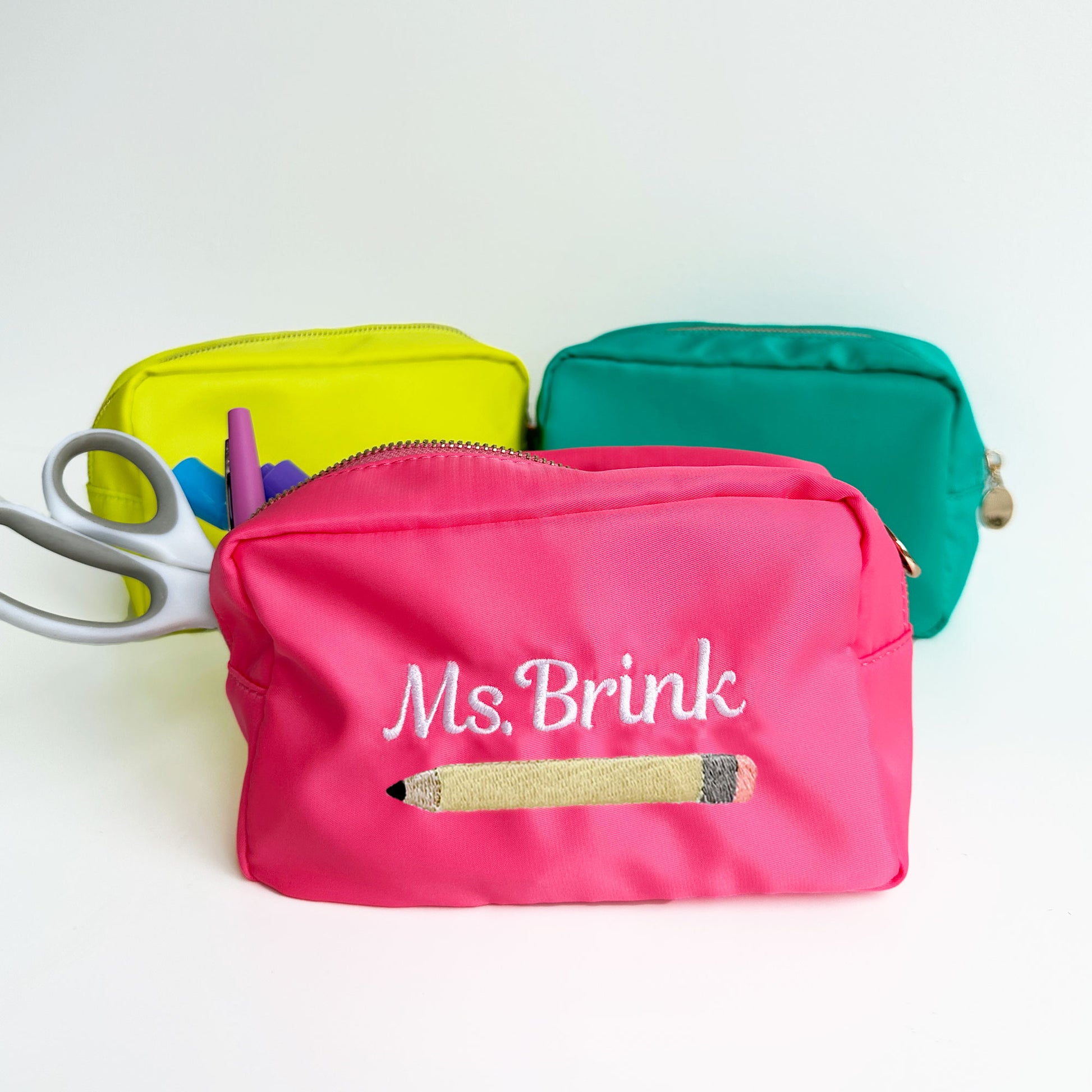 styled photo of a neon yellow, turquoise, and coral pink pencil bag pouches with emrboidered ms brink and pencil design on the coral pink bag. 