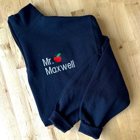 image of a navy blue quarter zip with a custom  embroidery design of a name with a mini apple