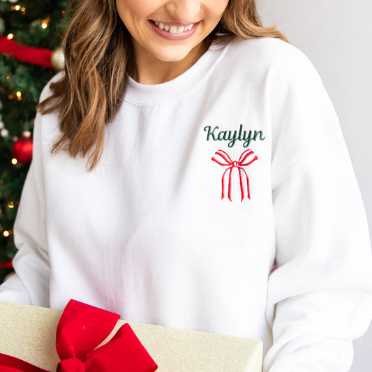 woman holding a wrapped christmas present near her tree wearing a white crewneck sweatshirt with her name and a festive bow embroidered on the left chest in green, red, and white