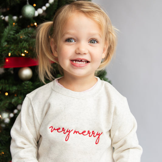 toddler girl wearing a heather oatmeal crewneck with very merry embroidered across the chest in a script font and red thread