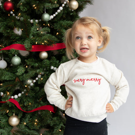 little girl standing in front of a christmas tree wearing an oat colored crewneck sweatshirt with very merry embroidered in a script font across the chest